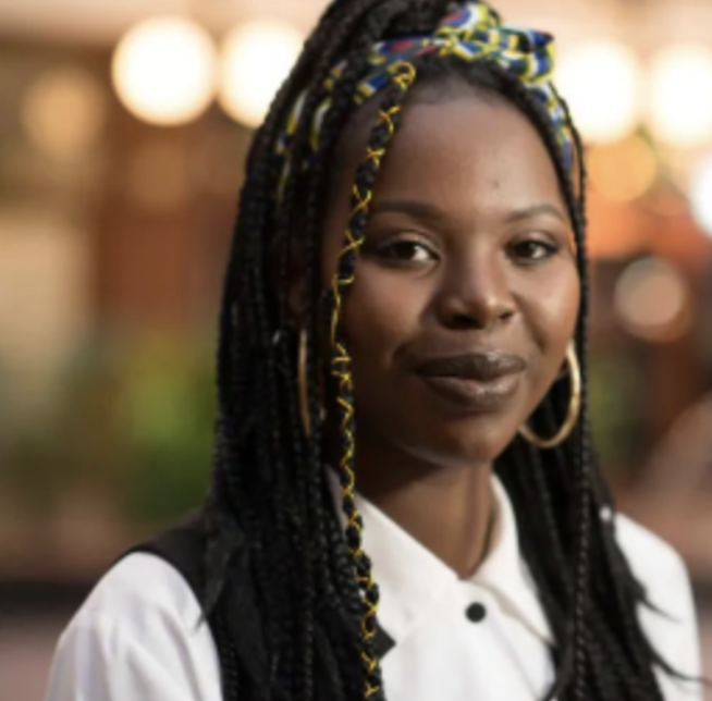 This photo is a portrait of Hasina, a dark-skinned Black woman. The background is blurry. She is at the forefront wearing a black vest on top of a white blouse. Her hair is in armpit-length box braids. It is in a half-up half-down style, tied together by a white, yellow, blue, and red Ankara scarf. The box braid framing the left side of her face is wrapped in gold thread. She is also wearing neutral everyday makeup and medium size gold hoops. She is smiling (without teeth) toward the camera.