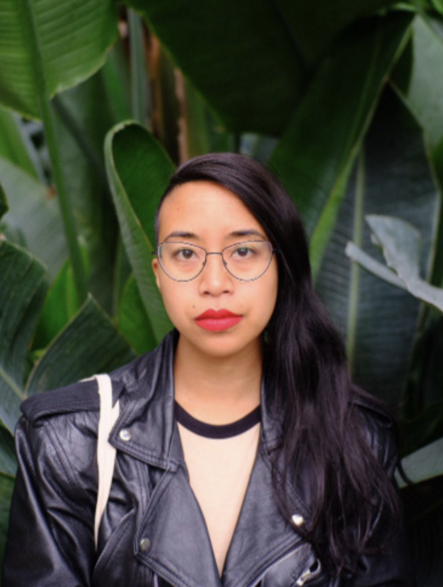 Attached is a photo of a Queer Non-Binary Filipino with partly shaved head and long hair on the side, looking at the photographer. The person is wearing black metal cat eye shaped glasses, red bold lipstick, a black leather jacket overtop a cream colored t-shirt with a black neckline trim. In the background is a forest of tall green banana leaves. 