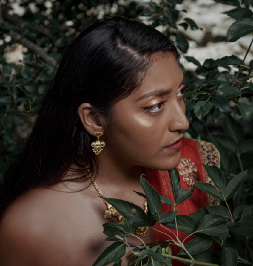 Amanda apparaît de profil dans un décor de verdure. Elle porte un sari rouge royal brodé de fil doré qui s’agence avec son rouge à lèvres rouge, ses traits de crayon à paupière noir et rouge, son ombrage à paupière rose doré et ses bijoux or. 